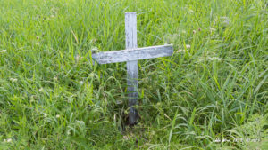 Dilapidated roadside memorial cross in long grass.